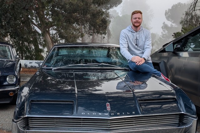 Man sitting on the driver side hood of his navy blue Toronado car